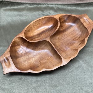 Könnte beinhalten: Ein Holzserviertablett mit drei Fächern und einer blattartigen Form. Das Tablett besteht aus hellbraunem Holz mit einer glatten, polierten Oberfläche. Die Fächer sind oval und blütenblattförmig, ideal zum Servieren von Snacks oder Vorspeisen.