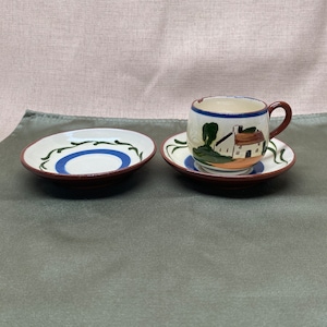 Puede incluir: Un juego de dos tazas de té de cerámica blanca con diseños florales azules y verdes. La taza de té y el platillo tienen una escena de cabaña pintada.