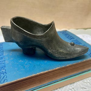May include: A small, antique-style metal shoe figurine with a high heel and a rounded toe. The shoe is a dark, muted gray color and has a small decorative ball on the toe. It sits on a blue book.