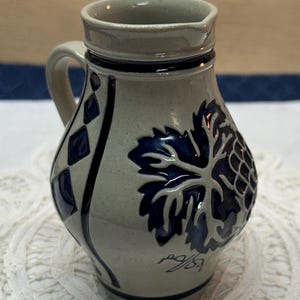 May include: A stoneware pitcher with a handle, featuring a blue grape leaf and cluster design. The pitcher has a light gray base color with dark blue accents and a dark blue rim. The pitcher is approximately 6 inches tall.