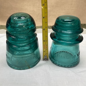 May include: Two antique teal glass insulators, one marked "Patented Oct 1907" and the other "Made in USA". A ruler shows the insulators are approximately 7.6 cm tall. These vintage pieces have a classic design and are likely from a telegraph or telephone system.