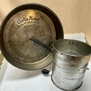 Pode incluir: Uma peneira Bake King e uma peneira de medição Bromwell's vintage. A peneira Bake King é uma peneira grande, redonda, de metal com uma alça e o texto "Bake King King of Bakeware Made in U.S.A." A peneira Bromwell's é uma peneira de medição de metal.