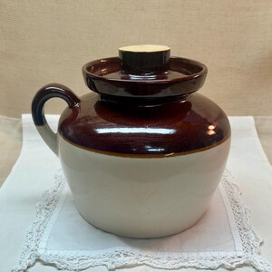 Op de afbeelding: Een keramische pot met een rond lichaam, voorzien van een glanzend donkerbruin glazuur op het bovenste gedeelte en een gladde crèmekleurige afwerking op het onderste deel. Het bevat een bijpassend donkerbruin deksel met een centrale knop en een handvat.