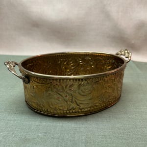 Peut inclure: Un bol ovale en laiton présentant un motif floral en relief sur toute sa surface extérieure. Le bol comprend deux petites poignées décoratives en métal argenté fixées sur les côtés opposés, contrastant avec le corps en laiton doré.
