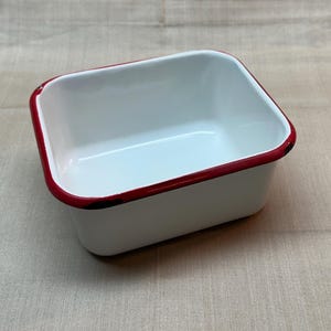 May include: A rectangular enamel basin with a glossy white interior and exterior finish. The container features a distinct, thin red rim along the top edge, providing a sharp color contrast against the white surface of the metal vessel.