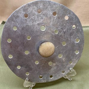 Könnte beinhalten: Ein kreisförmiges, silberfarbenes Metallobjekt mit zahlreichen, gleichmäßig verteilten Löchern. Es hat einen zentralen, hellfarbigen Holzknopf. Das Objekt wird auf einem transparenten Kunststoffständer präsentiert, was darauf hindeutet, dass es sich um einen dekorativen oder funktionalen Gegenstand handelt.