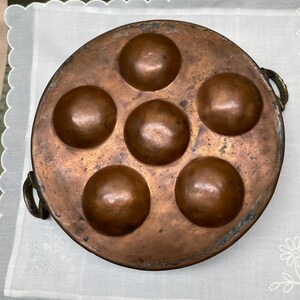 May include: A round, copper escargot pan with six recessed wells. The pan has a dark patina and two small handles. The wells are evenly spaced and have a rounded shape. The pan is designed for cooking and serving escargots.