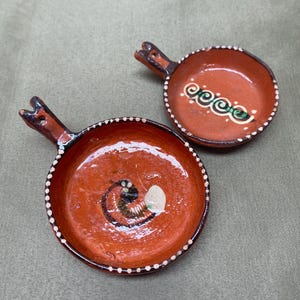 May include: Two small, terracotta-colored ceramic dishes with handles. Each dish features a hand-painted design in green, white, and black. The rims are decorated with small white dots. These dishes are ideal for serving dips or small portions.