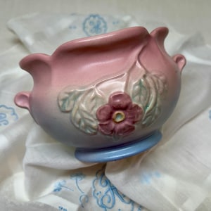 May include: A ceramic bowl featuring a gradient color scheme that transitions from a soft pink at the top to a light blue at the base. The exterior is decorated with a raised, molded floral motif consisting of a central pink flower and green leaves.