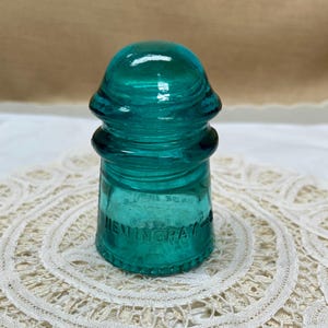 May include: A teal-colored Hemingray glass insulator. The glass has a rounded top and a cylindrical base with the word "HEMINGRAY" embossed on the side. The insulator is sitting on a white lace doily.