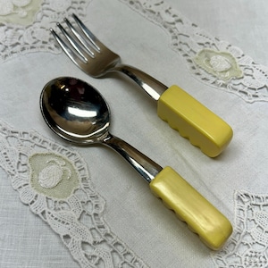 May include: A spoon and fork set with stainless steel heads and yellow handles. The handles have a ridged design for grip. The spoon has a rounded bowl, and the fork has four tines. The set is likely for a child or someone with limited mobility.
