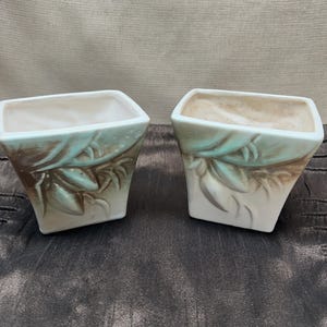 May include: Two square ceramic planters featuring a tapered base and a wide opening. The exterior surface is off-white with embossed leaf patterns highlighted by brown and soft green shading, creating a textured appearance on all visible sides.