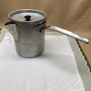 May include: A vintage aluminum coffee pot with a lid and a long handle. The pot has a spout and a black knob on the lid. It sits on a white cloth with a scalloped edge.