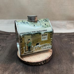 May include: A metal maple syrup container shaped like a log cabin with a silver screw-top lid. The exterior features a printed winter scene with a person, a cat, and a woodpile. Text reads Absolutely Pure Maple Syrup and 16.9 FL. OZ (1/2 L).