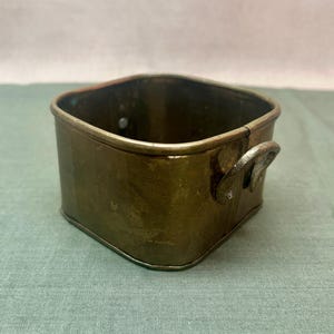 May include: A square-shaped brass container with rounded corners and a weathered, metallic finish. The vessel features two small, riveted metal handles on opposite sides and an open top, showcasing a dark, aged patina throughout the exterior.