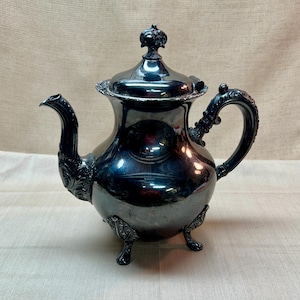 Op de afbeelding: Een gepolijste zilverkleurige theepot met een sierlijk handvat, een gebogen tuit met decoratieve details en een deksel met een knop. De pot staat op drie kleine, gedetailleerde pootjes en heeft een reflecterend, metalen oppervlak.