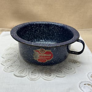 May include: A dark blue enamelware bowl with a white speckled pattern and a single side handle. The bowl features a red and gold label that reads Columbian Eneledware. The interior and exterior surfaces have a glossy finish with visible wear.