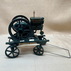 Op de afbeelding: Een donkergroen metalen model van een stationaire McCormick-Deering motor gemonteerd op een wagen met vier wielen. De motor is voorzien van een groot vliegwiel, een verticale uitlaatpijp en gouden letters met de tekst McCormick-Deering.