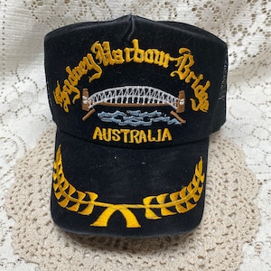 May include: Black trucker hat with gold embroidered text and design. The text reads "Sydney Harbour Bridge AUSTRALIA" above an image of the bridge. A gold laurel wreath design is on the brim.
