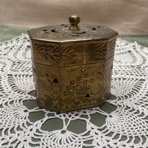 May include: A vintage brass hexagonal box with a lid and a small knob. The box is decorated with etched floral and grape designs. The lid has decorative cutouts. The box is sitting on a white lace doily.