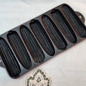 May include: A cast iron corn stick pan with seven corn-shaped molds. The pan is dark gray and has handles on each end. The molds are detailed to resemble ears of corn.