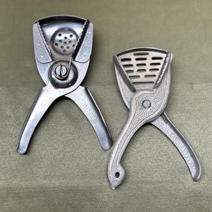 May include: Two vintage metal citrus juicers. One juicer has a perforated bowl and the other has a slotted bowl. Both have handles and a hinge mechanism. The juicer on the left has the text "VAUGHAN CHICAGO, ILL." on the handle.