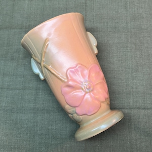 May include: A peach-colored ceramic vase with a large pink flower and leaf design. The vase has a flared top and a pedestal base. The flower is in relief, adding dimension to the design. The vase has a smooth, glossy finish.