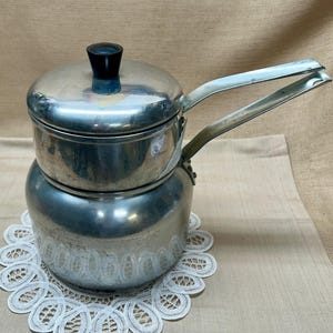 Puede incluir: Un juego de baño maría de acero inoxidable que incluye una olla de base redondeada y un recipiente insertable a juego con tapa. La tapa tiene un pequeño pomo negro y tanto el inserto como la olla inferior tienen mangos metálicos largos y planos.