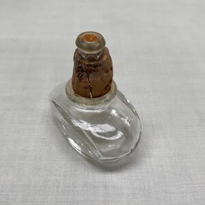 May include: A small, clear glass perfume bottle with an oval shape. The bottle has a cork stopper. The cork is brown and has a cracked texture. The bottle is sitting on a white surface.