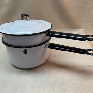May include: A white enamel double boiler set featuring two stacked pots with black rims and matching black handles. The top pot includes a fitted lid with a small handle. Both pots have a smooth white finish with black accents and circular holes at the ends of the handles for hanging storage.