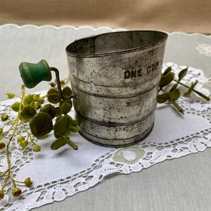 May include: A vintage metal measuring cup with a green wooden handle. The cup is marked "ONE CUP" and shows signs of wear. The cup is sitting on a white lace doily with green foliage.