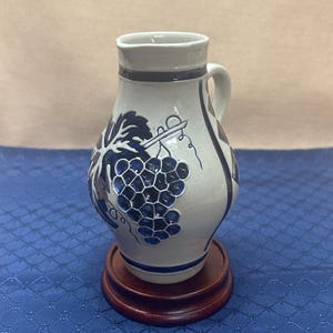 May include: A white ceramic pitcher with a dark blue grape design and handle. The pitcher sits on a round, dark wooden base. The pitcher has a dark blue rim and accents.