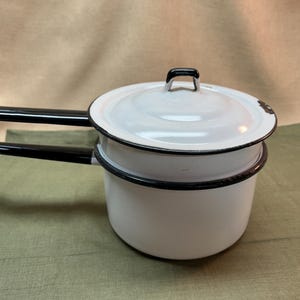 May include: A white enamel double boiler pot with a matching lid, featuring black rim detailing and two long black handles extending from the sides. The cookware has a smooth finish with a small area of dark wear visible on the lid edge.