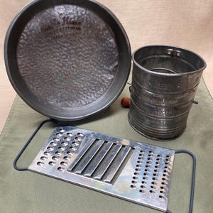 Pode incluir: Um conjunto de utensílios de cozinha vintage, incluindo uma forma redonda para assar com o texto "Bake King", uma peneira de farinha e um ralador de metal. Os itens são feitos de metal e mostram sinais de desgaste.