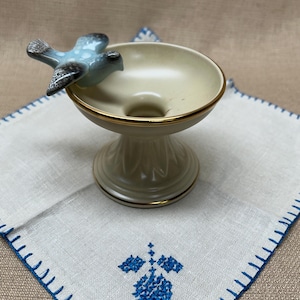 May include: A ceramic pedestal dish with a light blue bird figurine perched on the rim. The dish has a cream-colored interior and a gold rim. It sits on a white linen napkin with blue embroidered details.