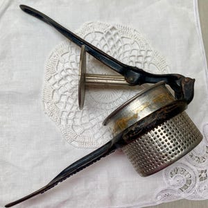 May include: An antique potato ricer with a black handle and a perforated metal bowl. The ricer has a lever mechanism and a circular pressing plate. The metal shows signs of age and use. The kitchen utensil is designed for mashing potatoes.