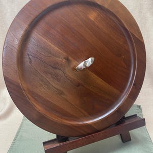 May include: A round, wooden serving tray with a dark brown finish. The tray has a raised edge and a small, silver-colored handle. The tray is displayed on a wooden stand.