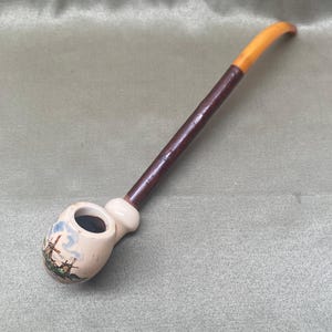May include: A vintage tobacco pipe with a cream-colored bowl featuring a landscape scene and a dark brown stem. The curved mouthpiece is a light amber color. The pipe is likely made of ceramic and wood.