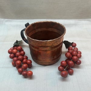May include: A cylindrical copper mug with a dark metal handle and a textured, weathered surface. The mug is flanked by two clusters of artificial red grapes with dark green leaves, creating a contrast between the metallic vessel and the fruit.