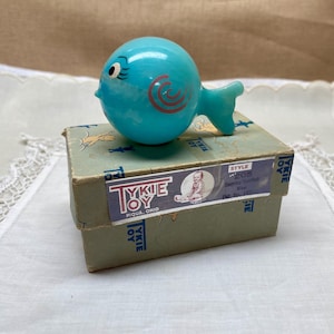 Puede incluir: Un juguete de pez vintage de Tykie Toy en color azul claro, con detalles en rojo y una cara sonriente. El juguete está sobre una caja vintage con el texto "TYKIE TOY PIQUA, OHIO" y "Sammy Sunfish".