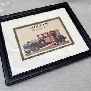 May include: Framed vintage Lincoln Motor Company advertisement. The black frame surrounds a print featuring a classic car and figures in period attire. The print has a white border and a green inner border. The Lincoln logo is visible at the top.