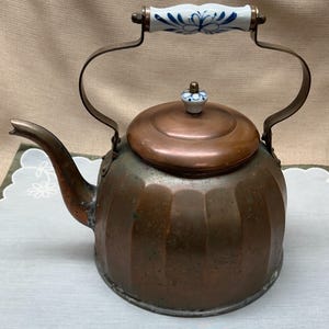 May include: A copper kettle with a round body and a curved spout. The lid and handle are copper-colored, and the handle has a white ceramic grip with blue floral designs. The kettle is designed for stovetop use.