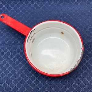 May include: A small, vintage saucepan with a bright red handle and rim. The interior is white, and the exterior shows signs of wear. The handle has a hole for hanging. This cooking pot is likely made of enamel-coated metal.