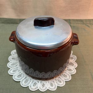 May include: A brown ceramic casserole dish with a silver lid and a dark brown handle. The dish sits on a white lace doily. The lid has a dark brown handle. The casserole dish is a kitchenware item.