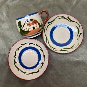 May include: A set of three ceramic teacups with a white background and blue and green floral designs. The teacup has a painted image of a white house with a brown roof and green trees.