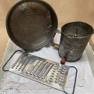 Pode incluir: Um conjunto de utensílios de cozinha vintage, incluindo uma forma de torta de metal com texto em relevo, uma peneira de farinha e um ralador. A forma de torta tem uma superfície texturizada. A peneira de farinha tem medidas marcadas na lateral. O ralador é retangular.