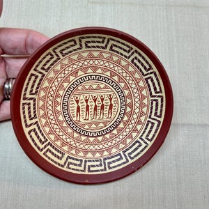 May include: A round terracotta-colored ceramic dish featuring a cream-colored interior with dark brown geometric patterns. The design includes a Greek key border, concentric triangles, and a central circular band depicting five stylized human figures.