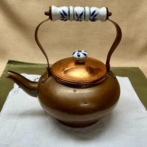 May include: A copper teapot with a matching lid and a decorative handle. The handle is made of white ceramic with blue floral designs. The lid has a small ceramic knob with a blue and white floral pattern. The teapot is sitting on a white cloth.