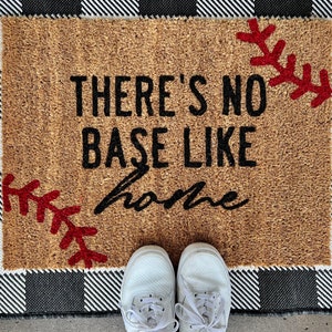 May include: A brown doormat with the text "There's no base like home" in black lettering. The doormat has red stitching details that resemble baseball stitches. The doormat is on a black and white checkered background.