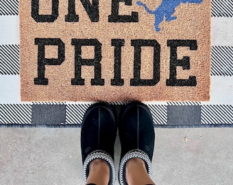 May include: A brown doormat with the words "ONE PRIDE" in black lettering and a blue lion graphic. A pair of black slippers with white trim are on a black and white checkered rug in front of the doormat.